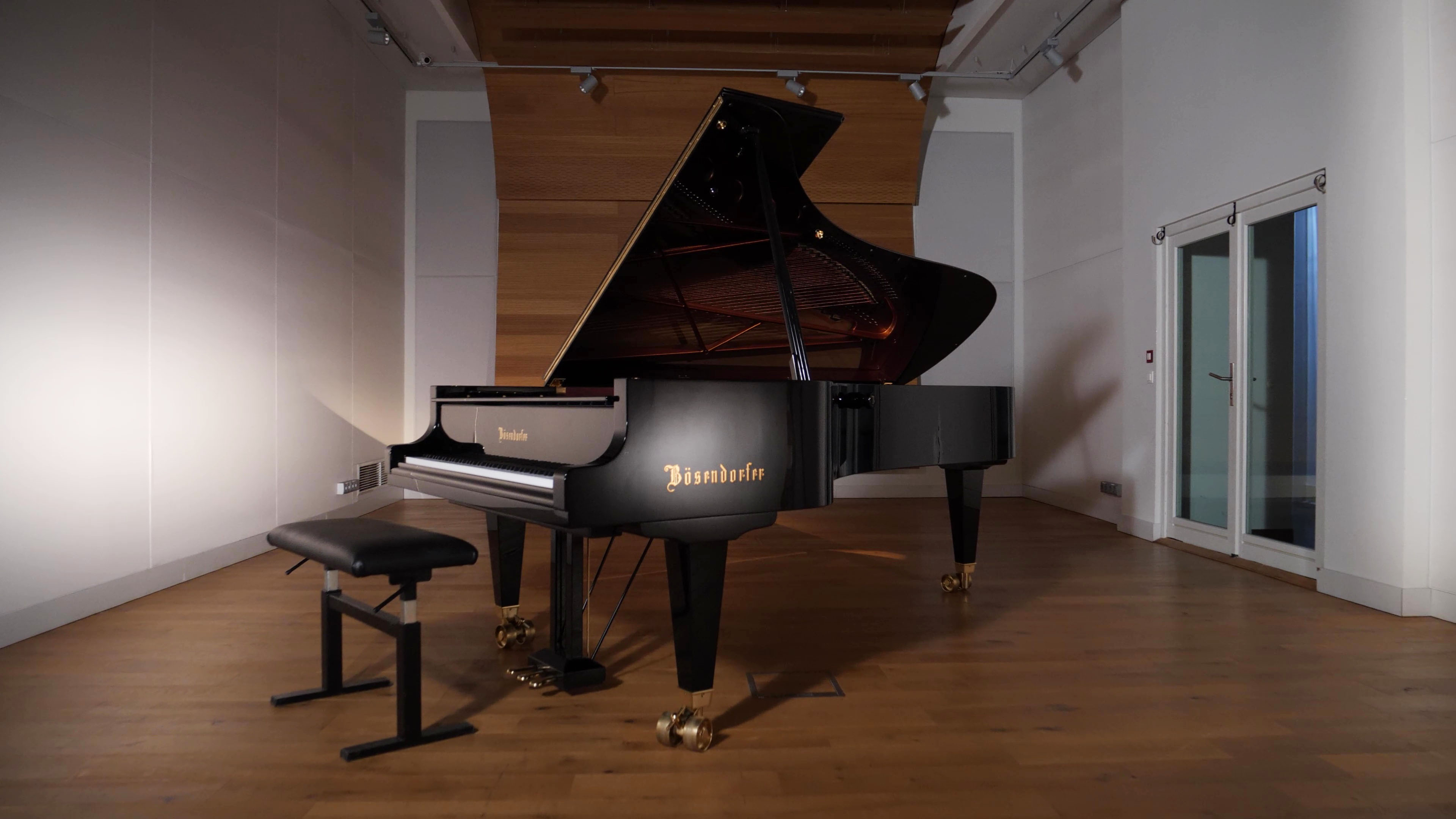 Full-size black Bosendorfer grand piano with its lid open, a matching black piano bench, and a music room with hardwood floors, white walls, and a wooden accent wall, and a door visible in the background.