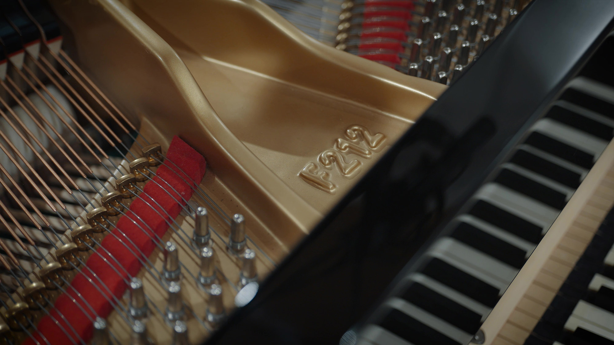 Close-up of a grand piano's interior showing strings, hammers, a red damper felt, part of the gold harp frame with F212 embossed, and black and white keys.