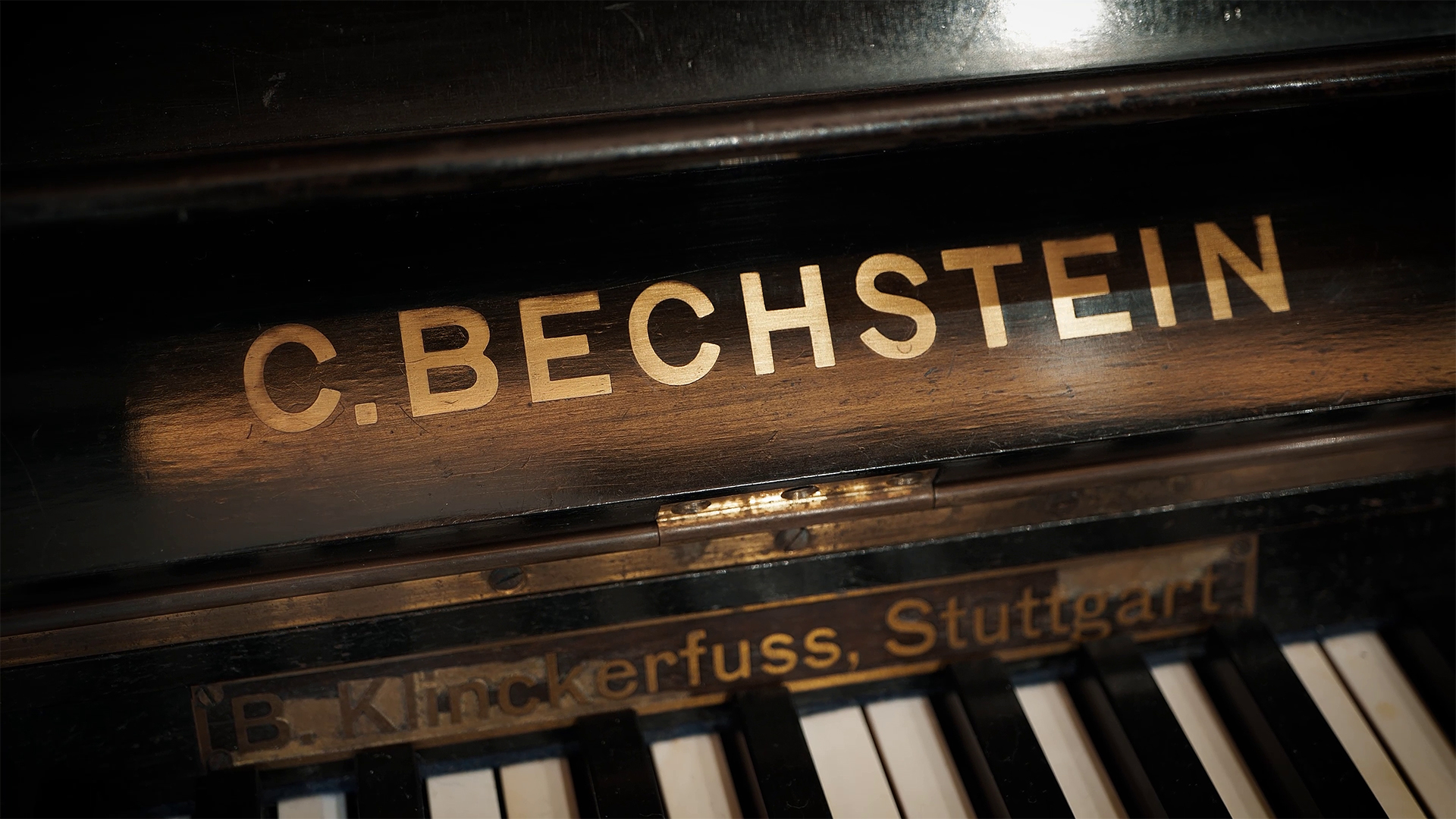 Close-up of a vintage black piano, showing the gold embossed 'C. BECHSTEIN' logo on the fallboard, with part of 'B. Klinckerfuss, Stuttgart' visible below and white and black piano keys in the foreground.