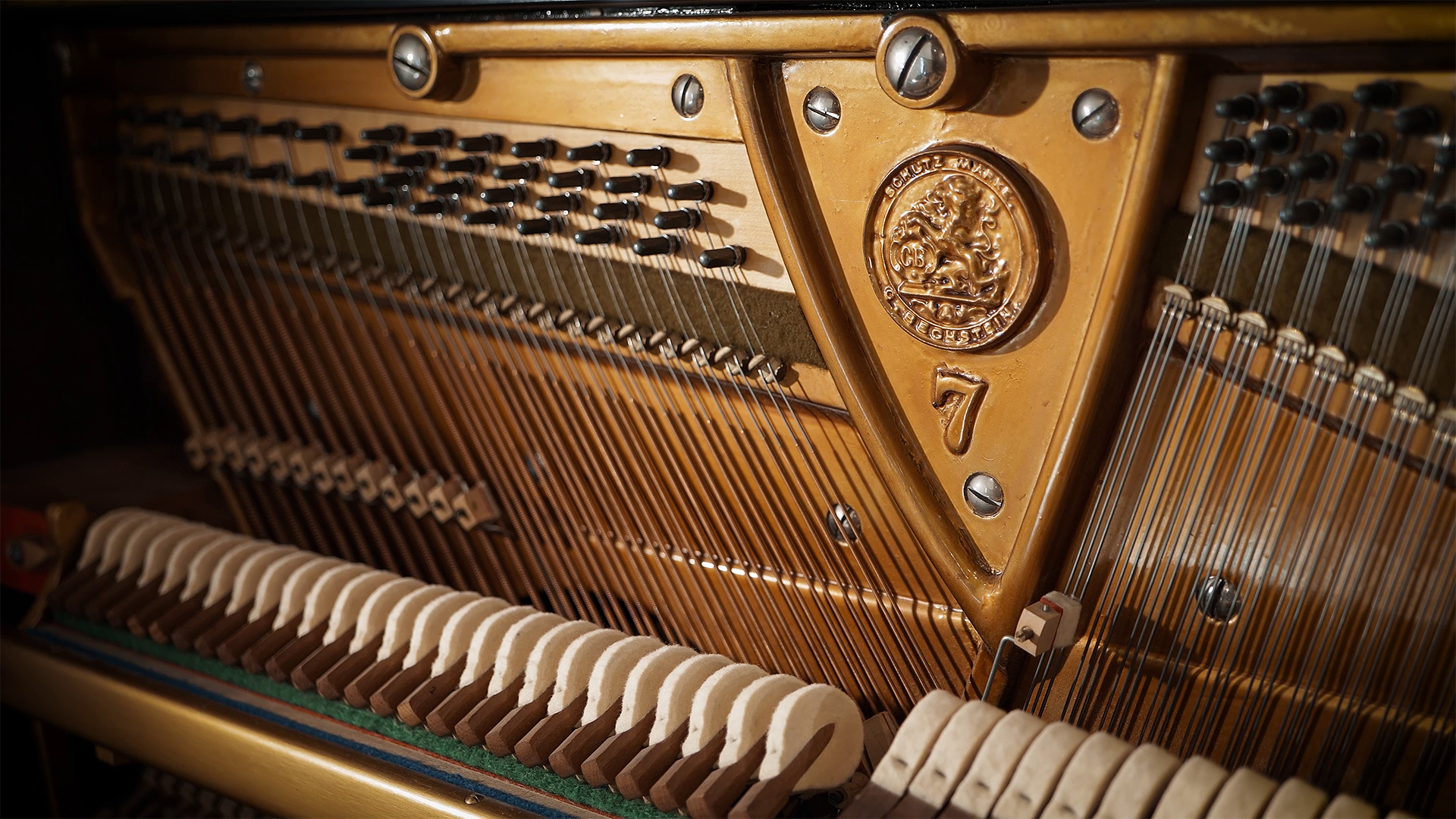 Close-up view of the intricate interior of an acoustic piano, showing the hammers, strings, and a bronze or gold-colored sounding board with a detailed medallion and the number 7.