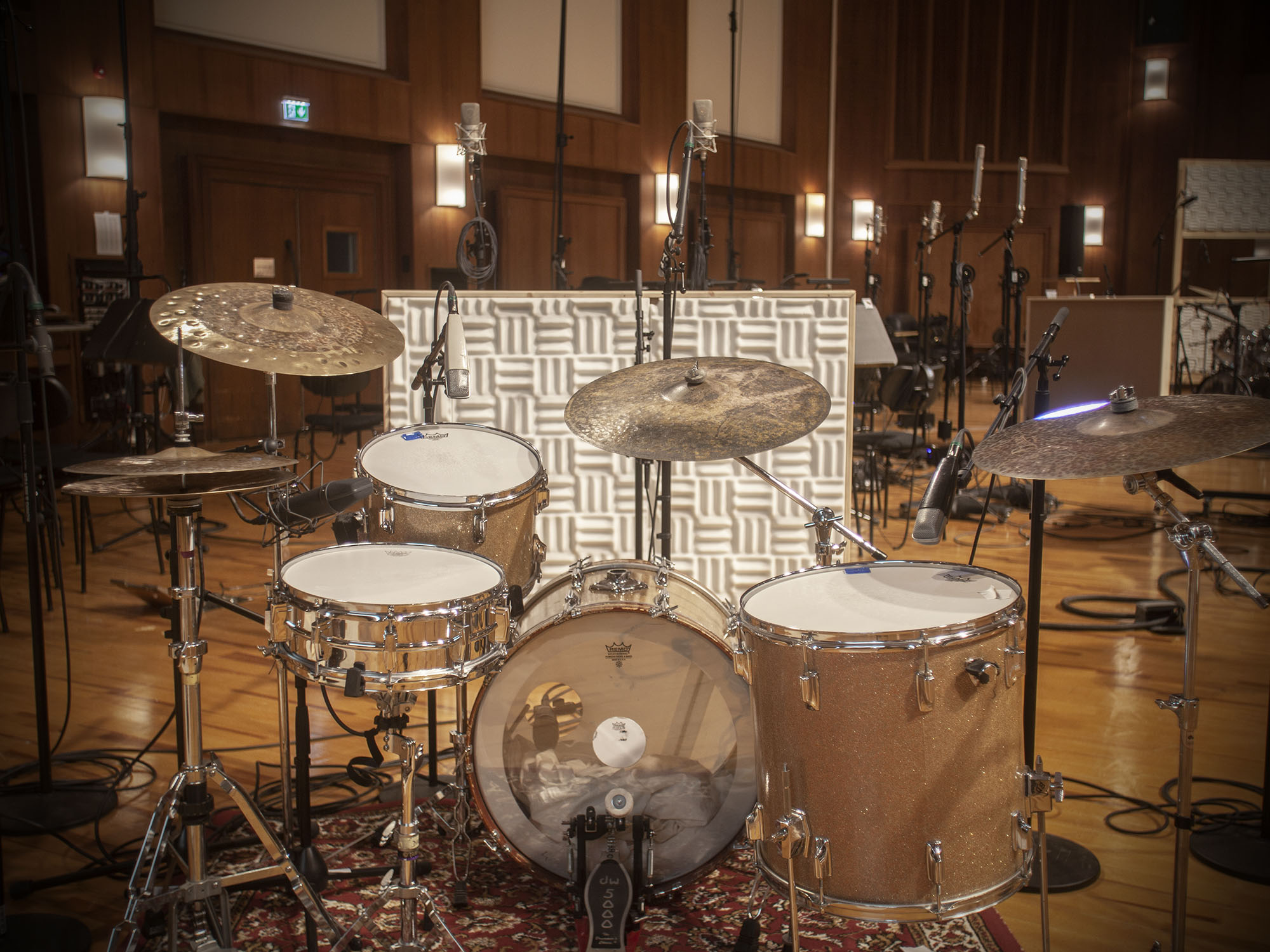 Full drum kit with cymbals and microphones, set up inside a professional recording studio with wood paneling and acoustic foam panels.