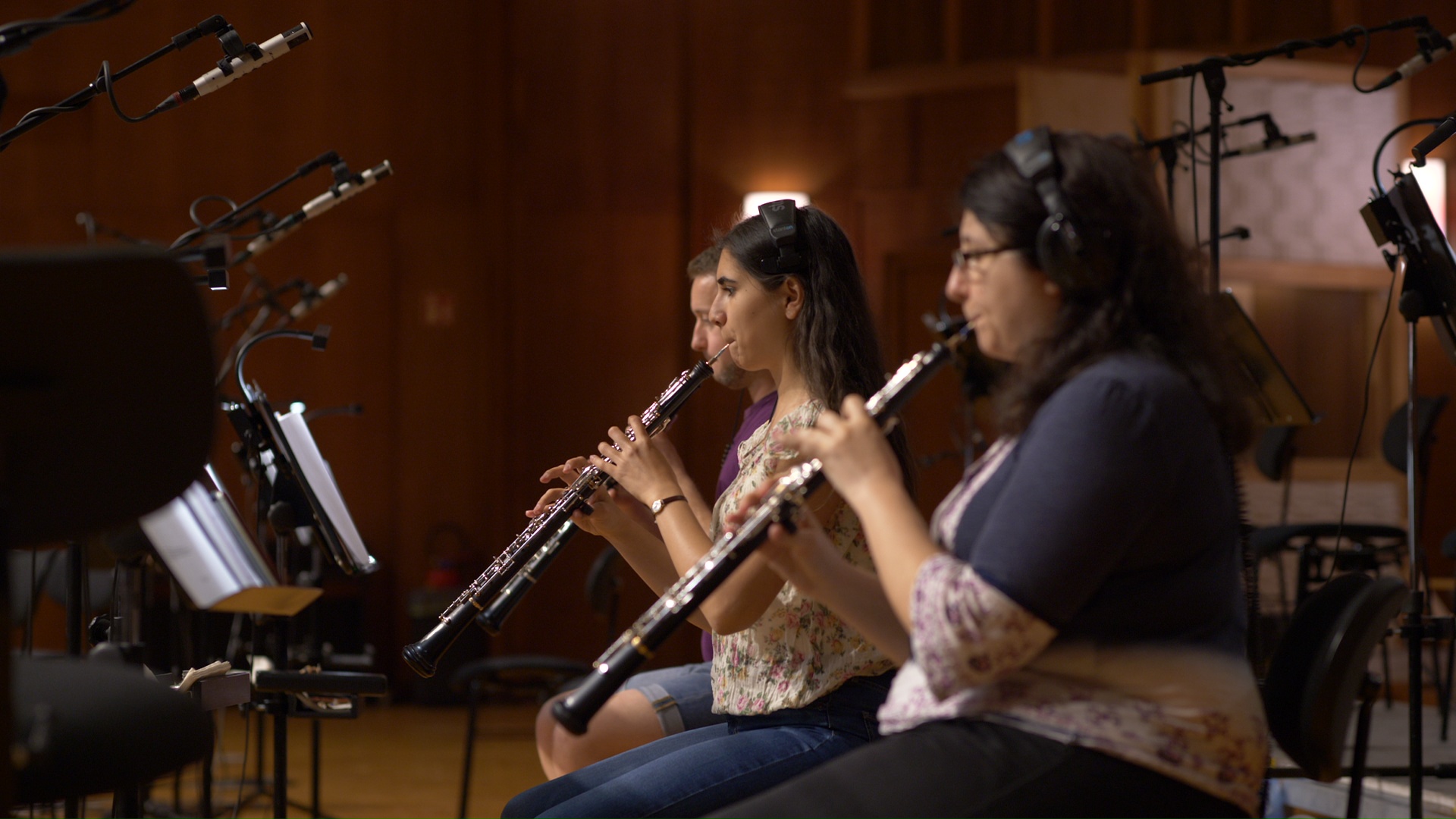 Musicians in headphones playing oboes in a recording studio, with microphones and music stands visible.