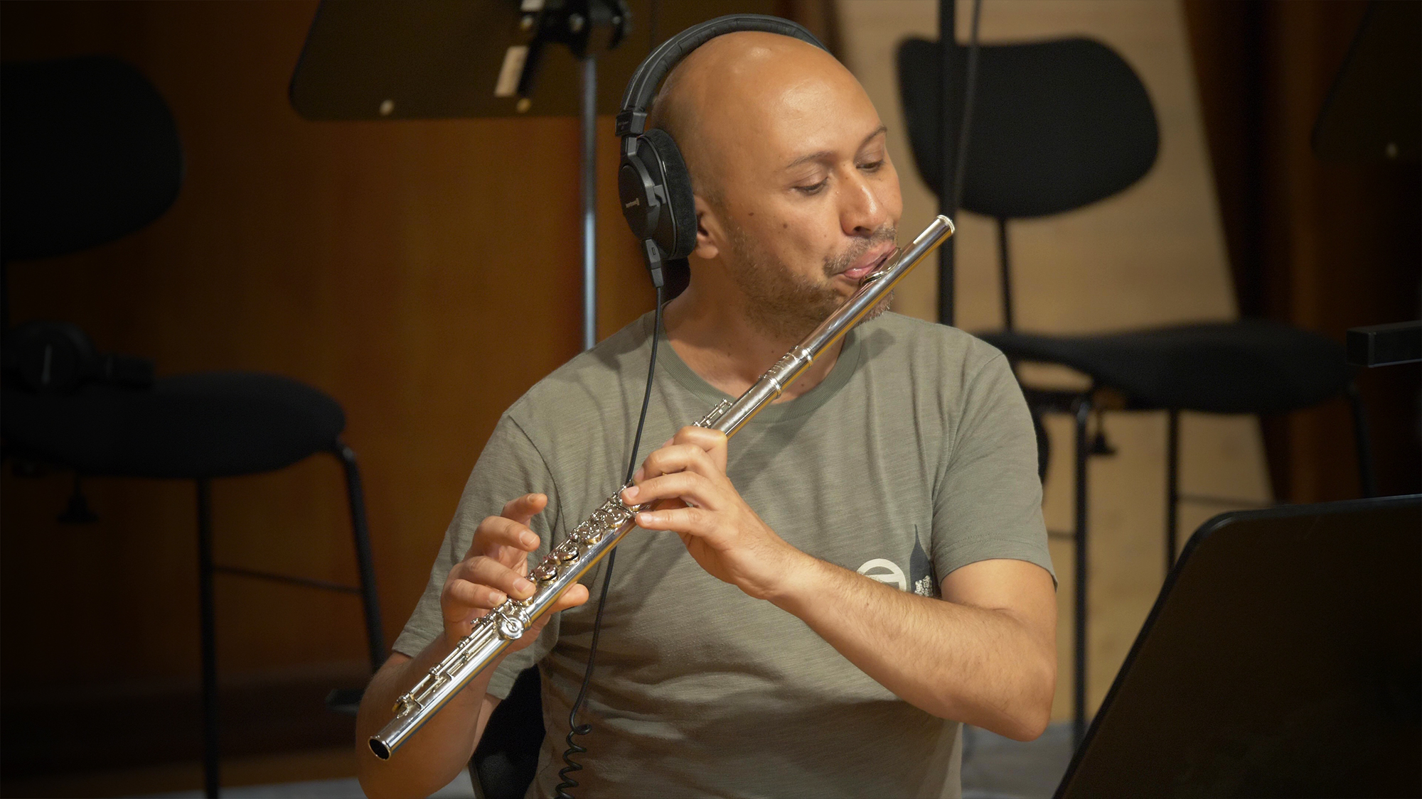 Bald man with headphones playing a silver flute in a recording studio.