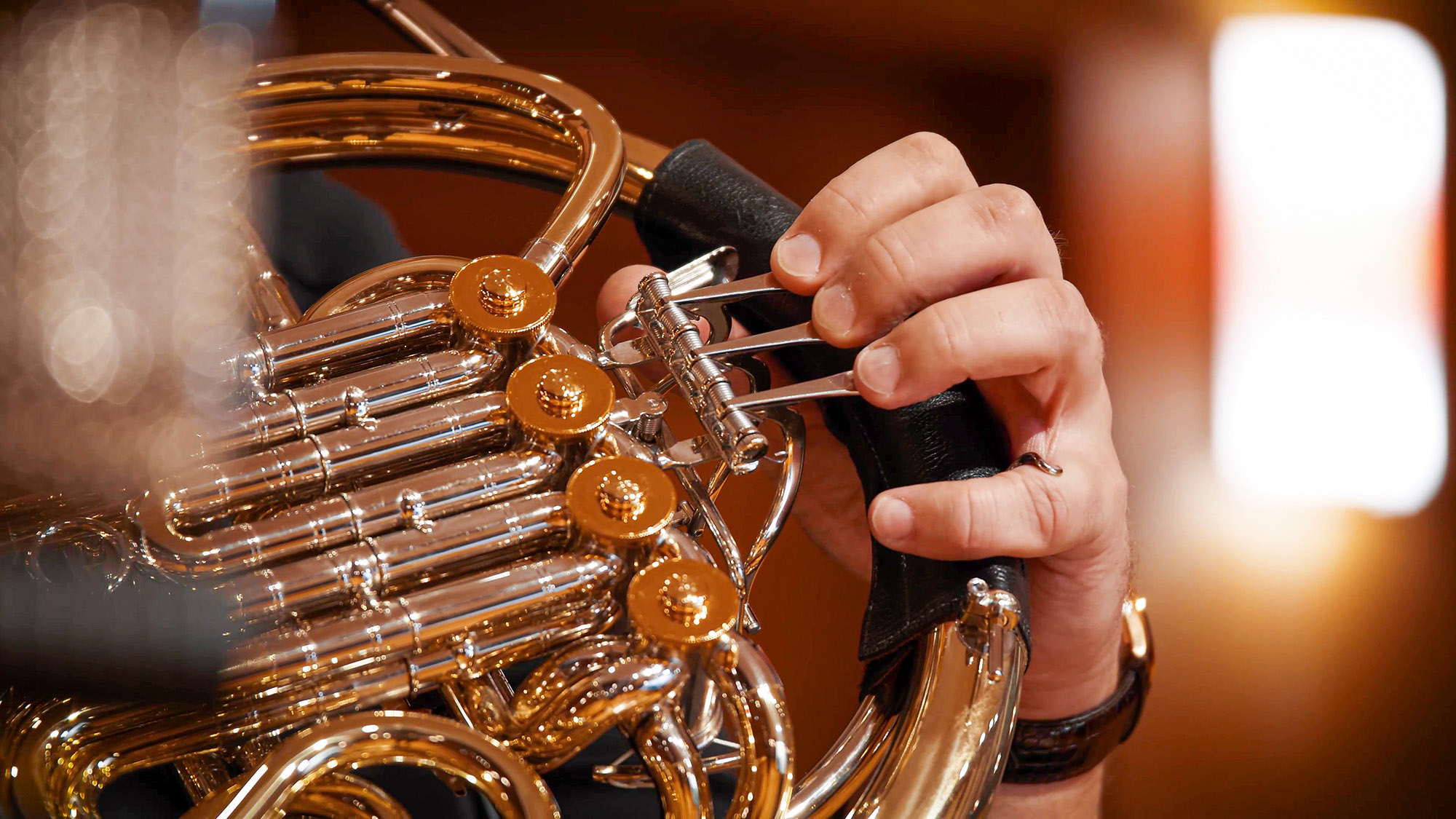Close-up of a musician's hand pressing valves on a golden French horn, highlighting the instrument's brass and chrome details.