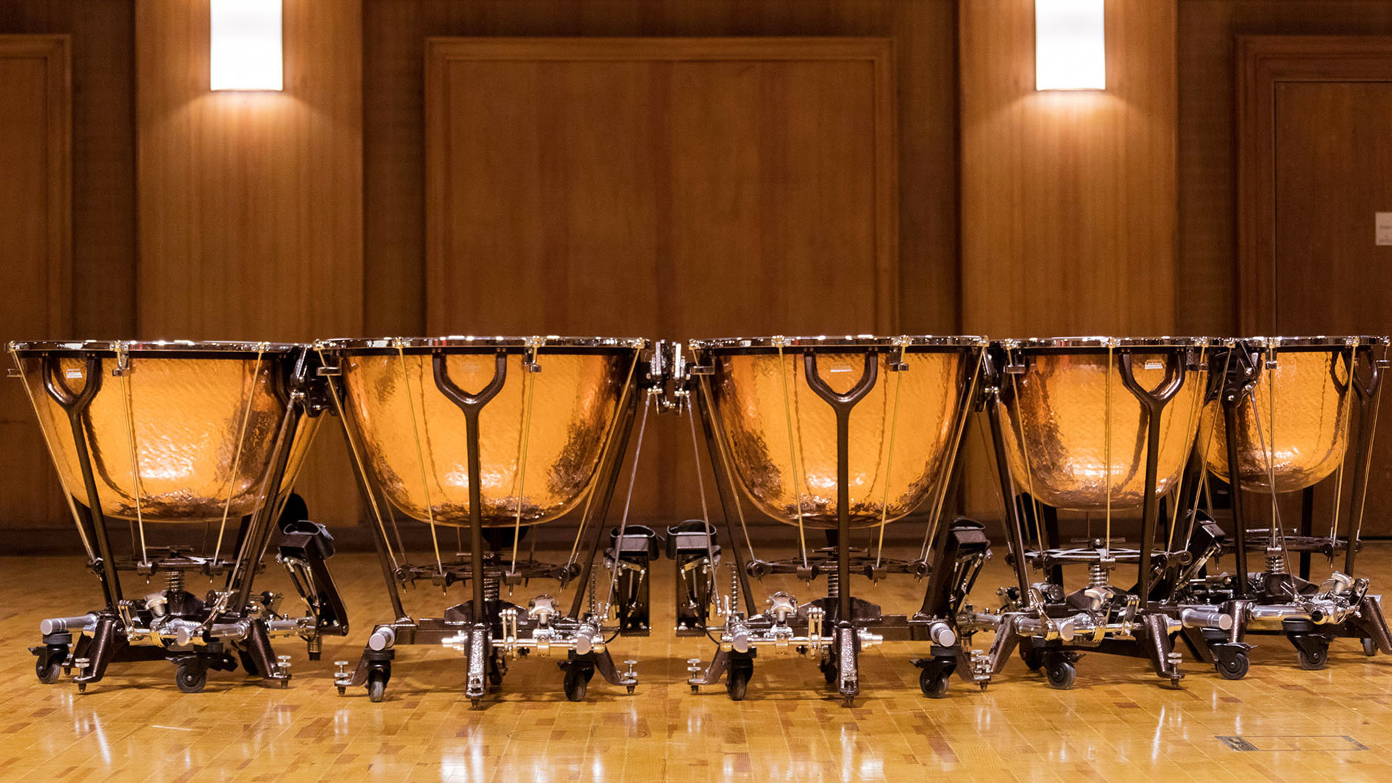 Four gleaming copper timpani with black stands and silver foot pedals arrayed on a polished wooden stage, illuminated by sconces.