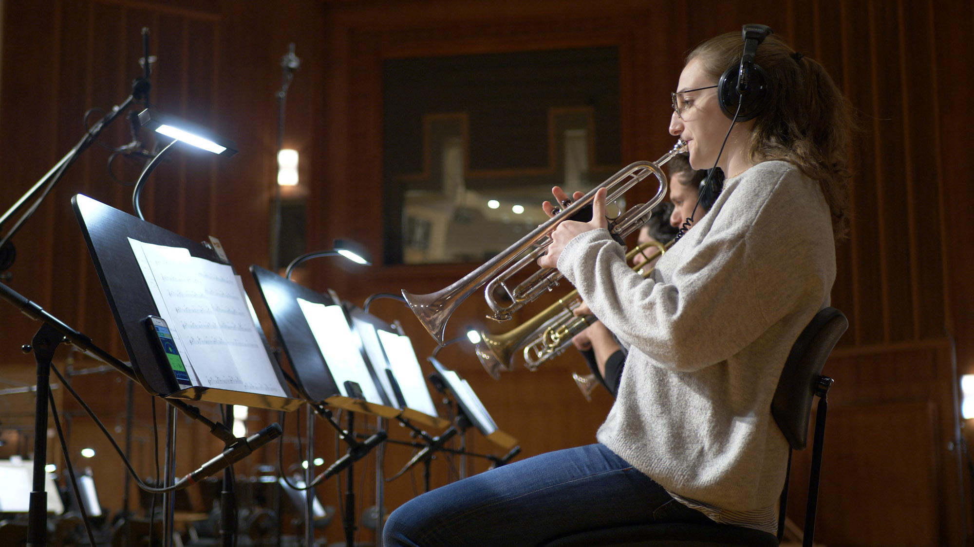 Young female trumpeter wearing headphones playing a silver trumpet in a recording studio with sheet music stands and bright lights.