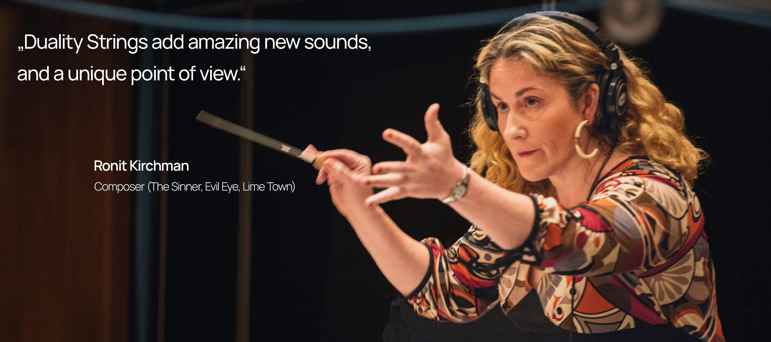 Composer Ronit Kirchman wearing headphones and patterned top, holding a conducting baton with one hand and gesturing with the other, standing in a large recording studio with dark background.