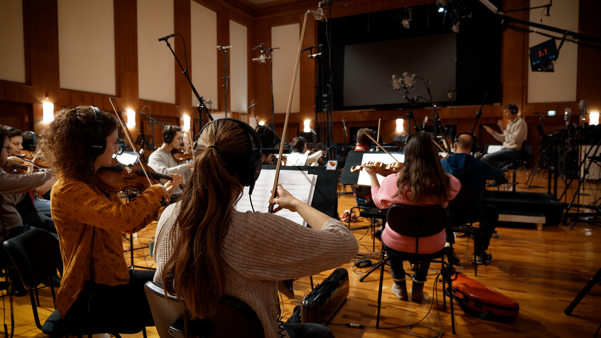 Full orchestra rehearsing or recording in a studio, musicians playing violins with headphones, microphones capturing sound.