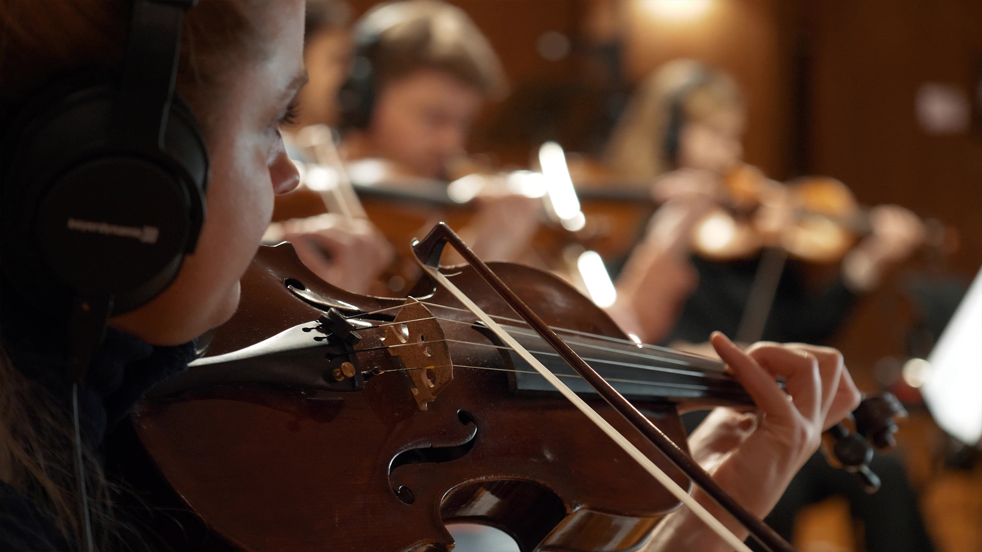 Close-up of a female musician with headphones playing a violin in an orchestra, surrounded by other musicians.