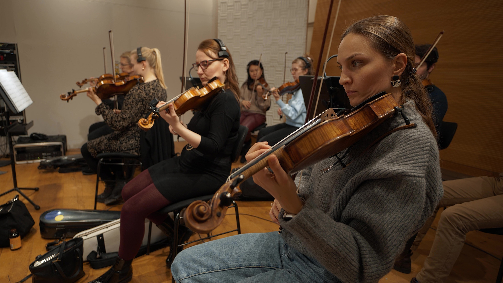 String musicians rehearsing in a recording studio, featuring multiple violinists and a cellist playing instruments, with music stands and cases visible.