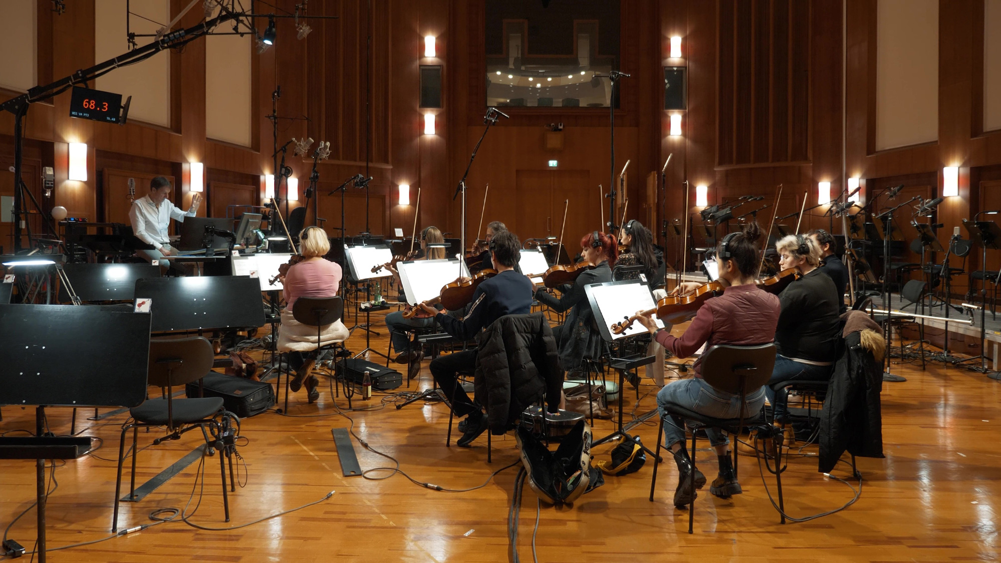 Full orchestra recording session in a professional studio, with musicians playing violins and other string instruments, and a sound engineer at the control desk.