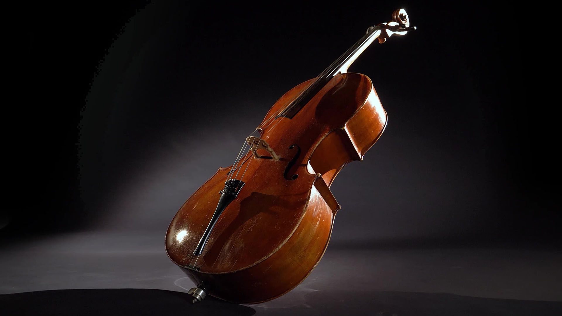 Brown wood cello standing on its endpin with strings and scroll visible, against a dark background with dramatic lighting highlighting the instrument's contours.