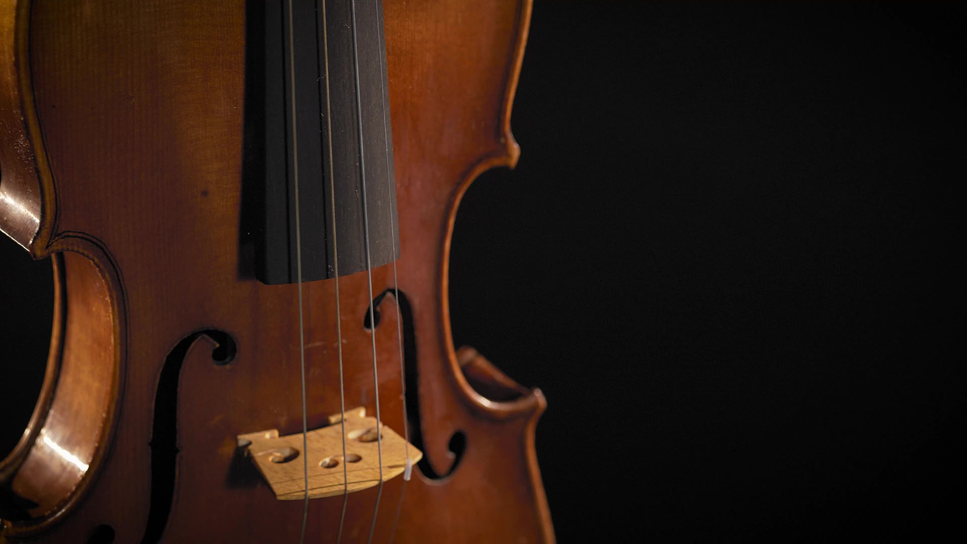 Close-up of a rustic cello or bass against a dark background, showing its warm wooden body, f-holes, strings, and bridge, highlighting the instrument's classic craftsmanship
