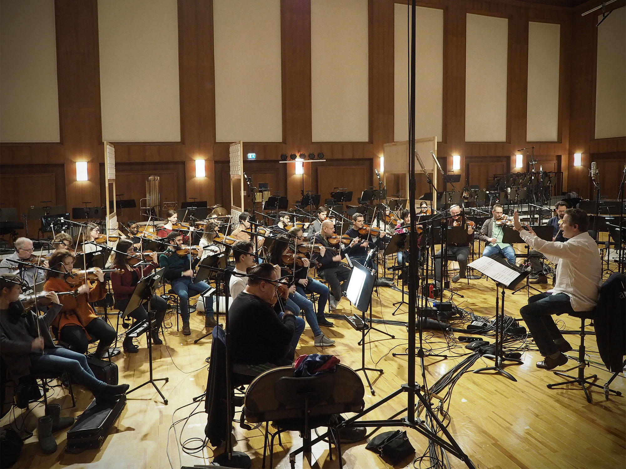 Professional orchestra performs in a recording studio, with musicians playing violins and other string instruments, surrounded by recording equipment and microphones.