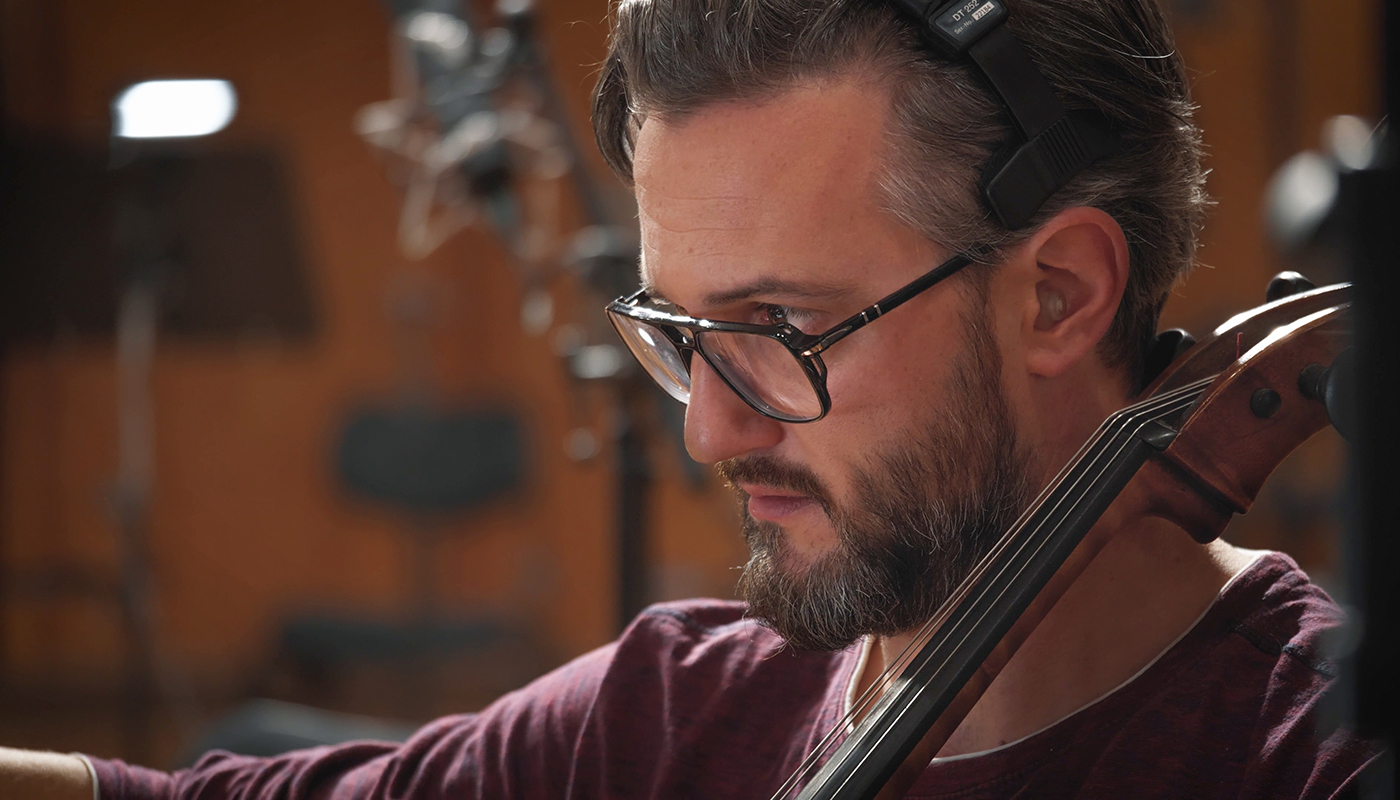 Musician with glasses and beard wearing headphones playing cello in a recording studio.