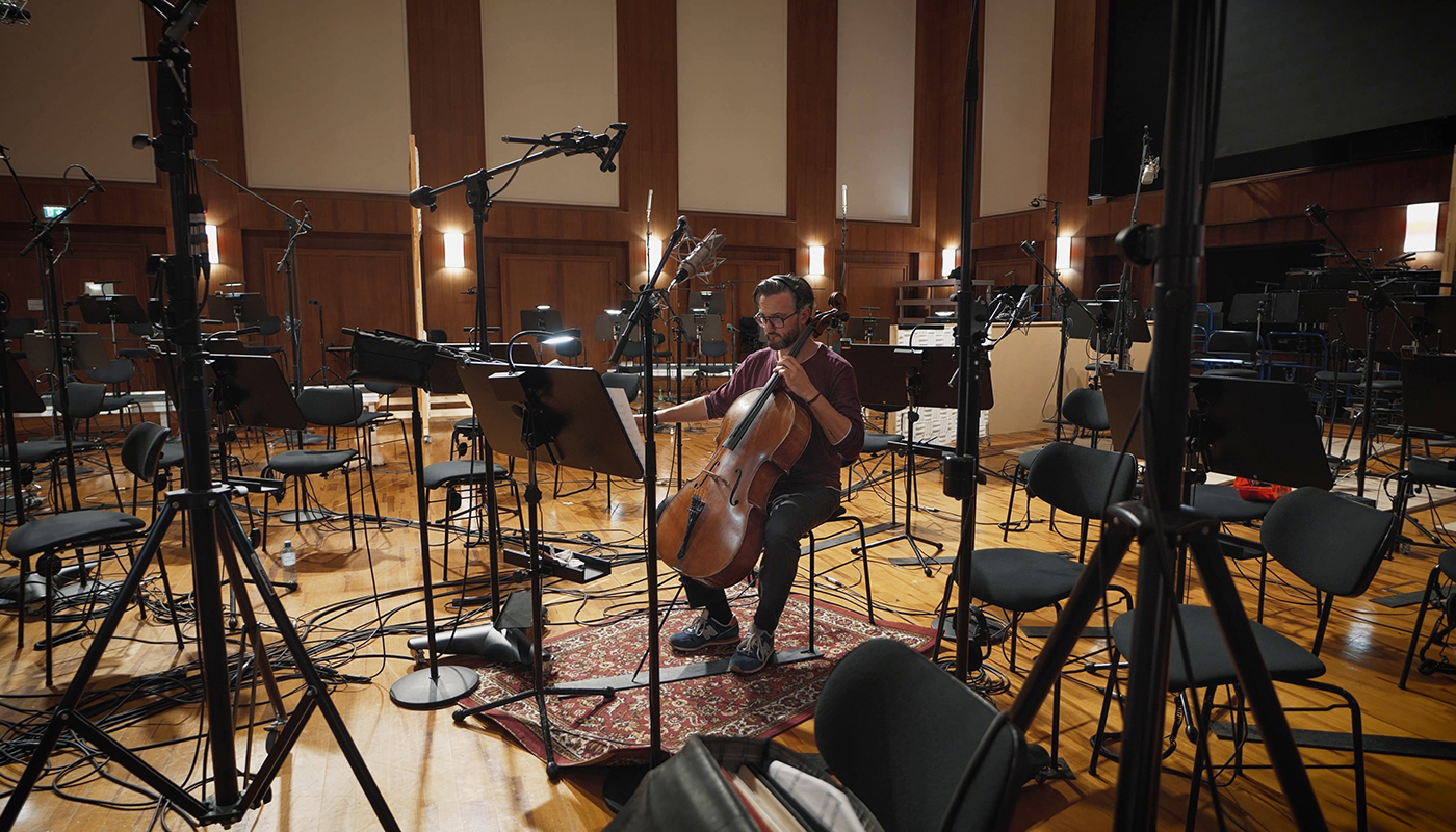Musician playing cello in a professional recording studio, surrounded by microphones, music stands, and chairs, with wood floors and paneling.