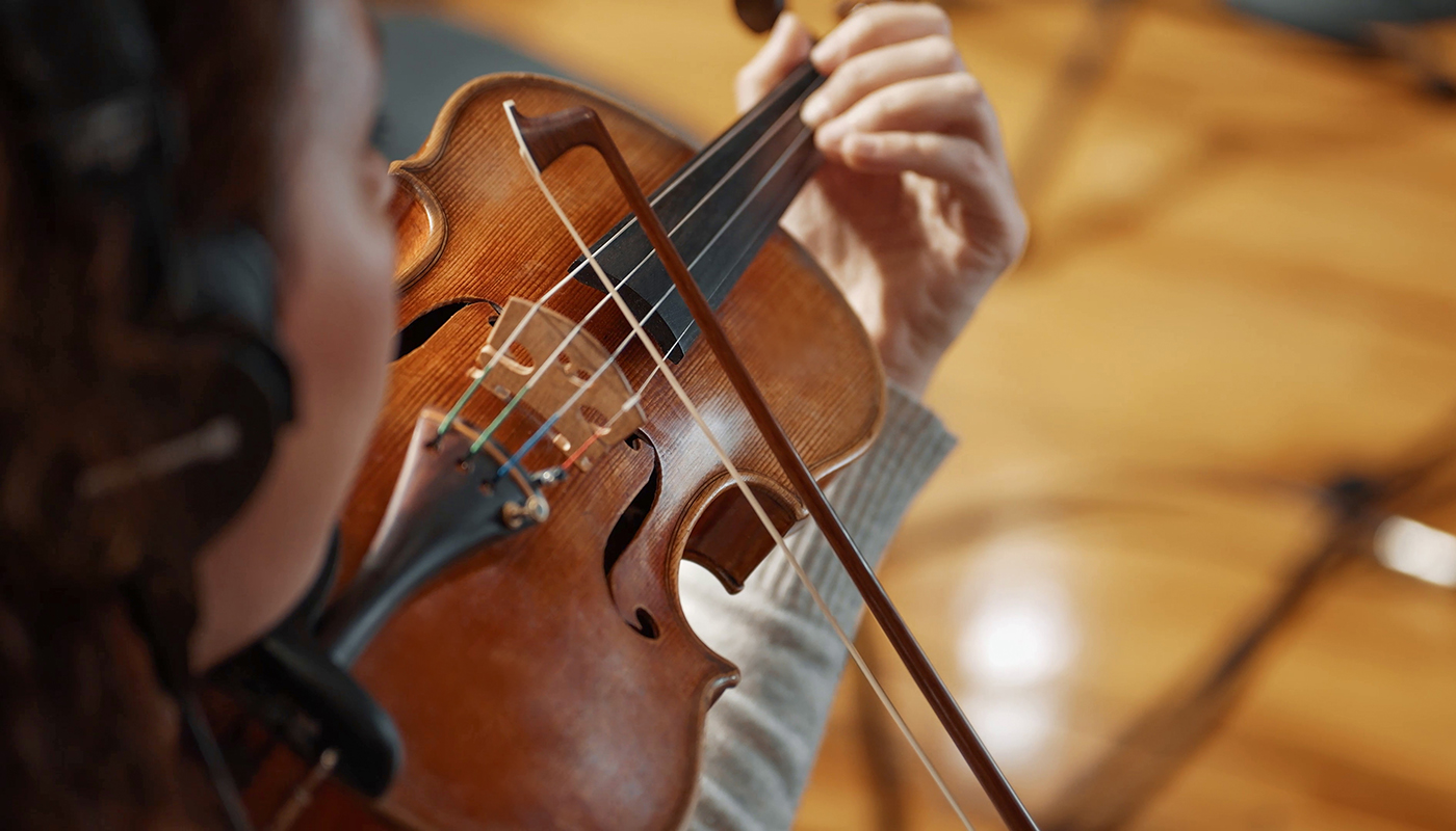 Musician wearing headphones playing a wooden violin with a bow, close-up view of the instrument's strings and bridge.