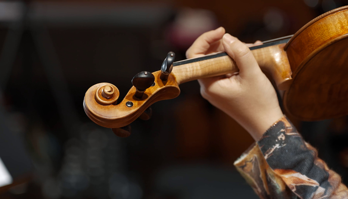 Close-up of a person's hand holding and playing a wooden violin, showing the scroll, pegbox, and tuning pegs.