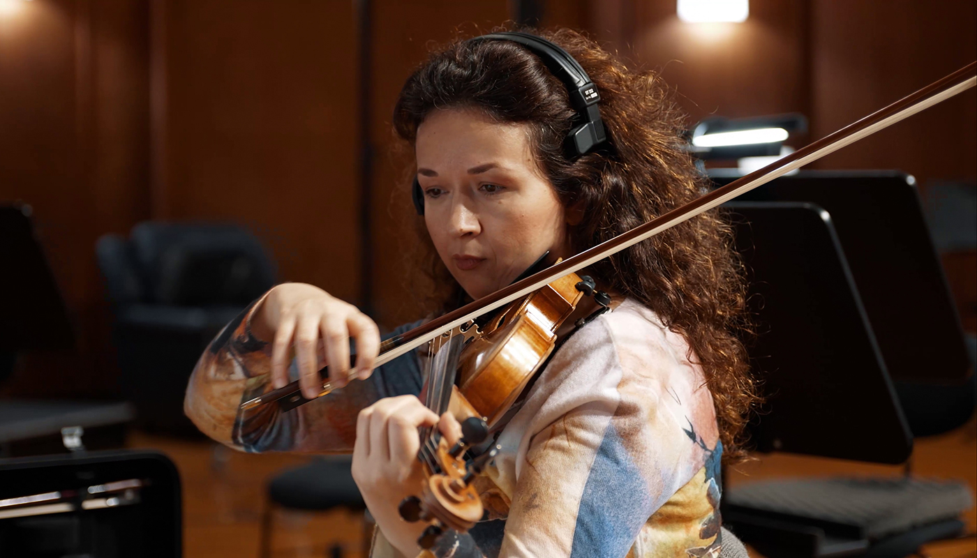 Woman with curly brown hair wearing headphones and a colorful top plays the violin with a bow in a recording studio, focused on her instrument.