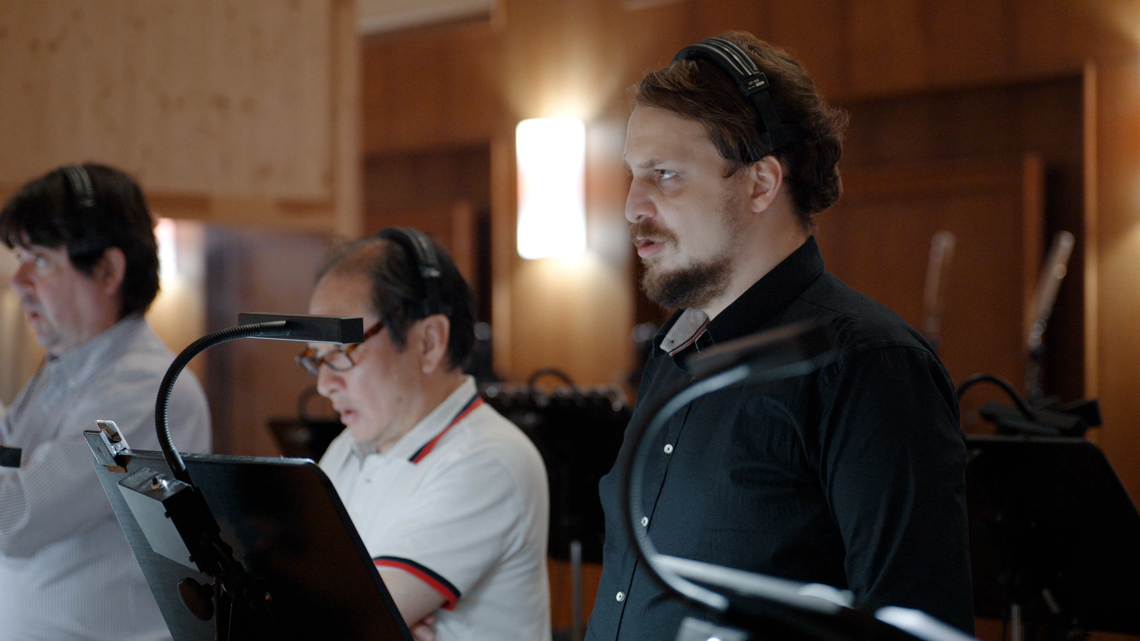 Three men with headphones in a recording studio, two looking at sheets on stands with task lights, one with a beard and black shirt turned right.