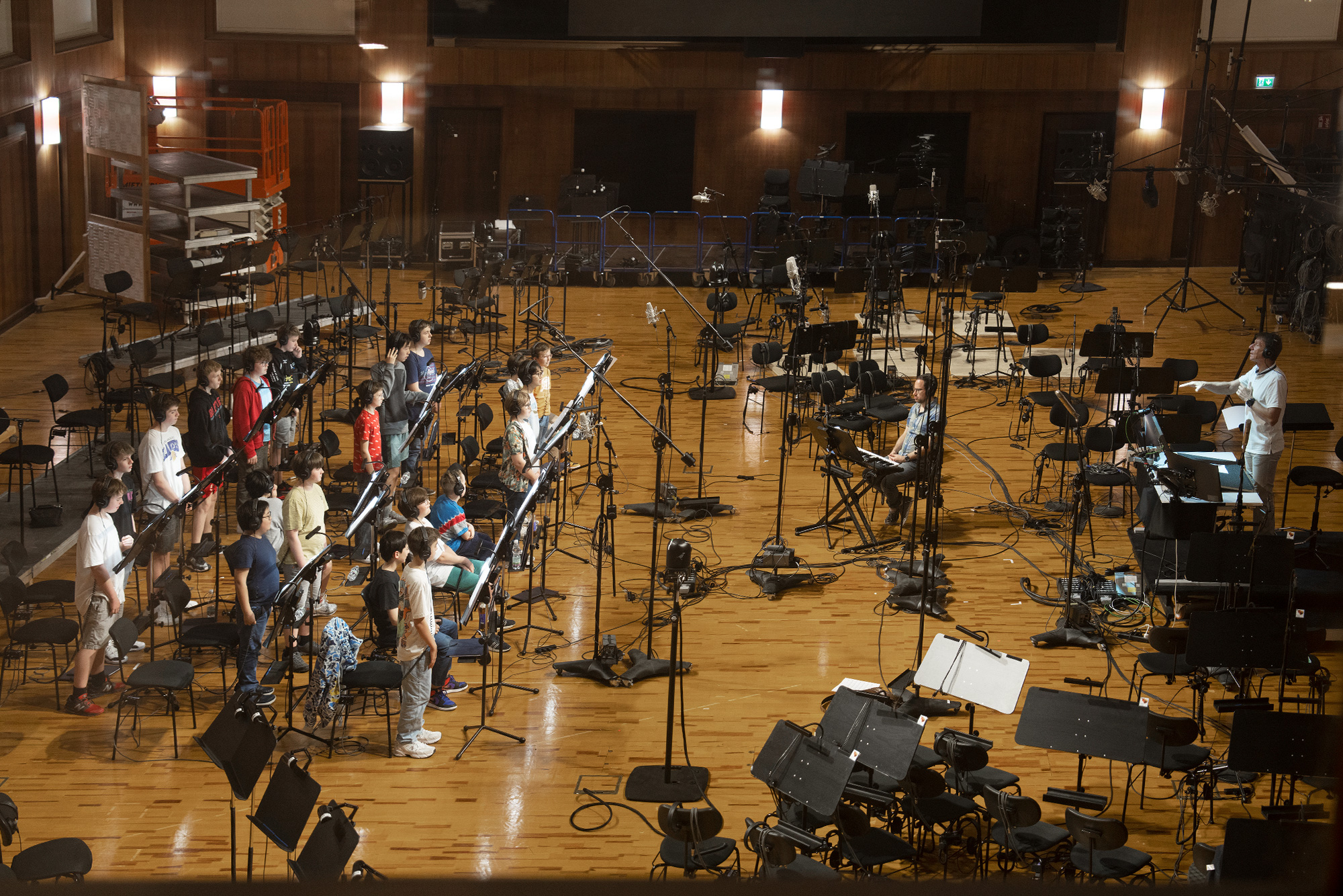 Large orchestral recording studio with young musicians and adults seated among music stands, microphones, and audio equipment on a wooden floor, with a director overseeing production.