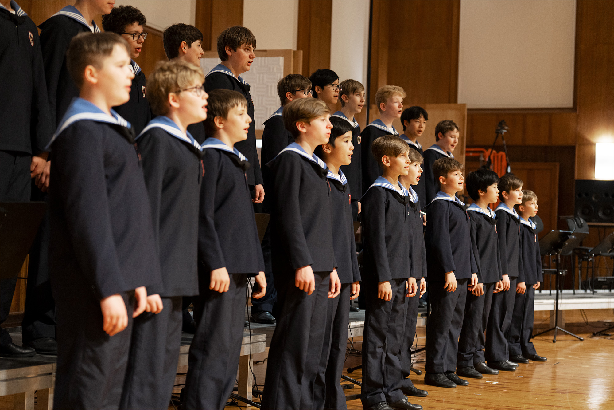 A diverse group of young boys in matching dark navy uniforms with white and blue sailor collars standing in rows on a polished wooden stage, singing with open mouths.