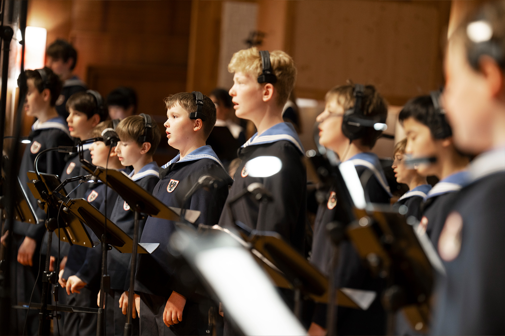 Young male choir members wearing navy and white sailor-style uniforms with headphones and microphones, singing or performing.