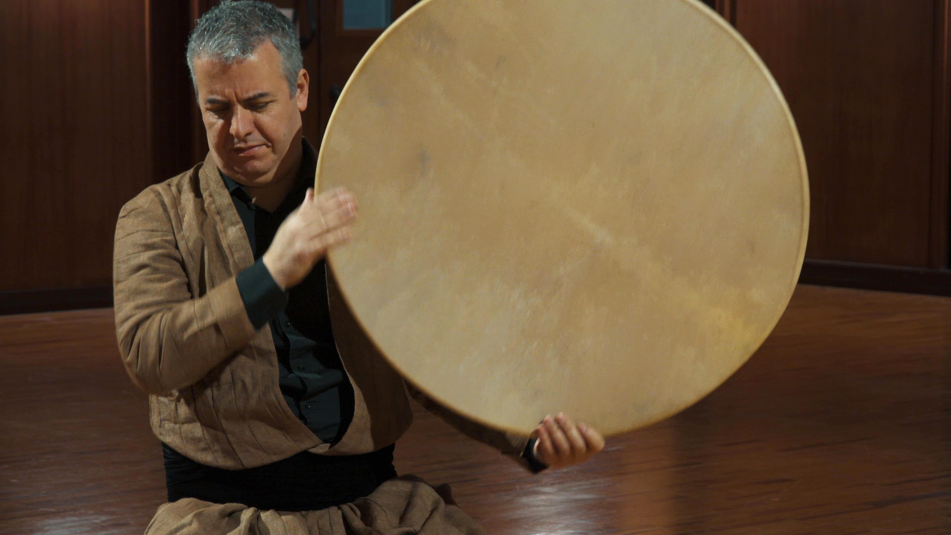 Man playing a large circular frame drum, possibly a daf or bendir, using his right hand to rhythmically strike the light-colored drumhead. He is wearing a traditional brown jacket and black shirt while seated on a wooden floor in a room with dark wood accents.