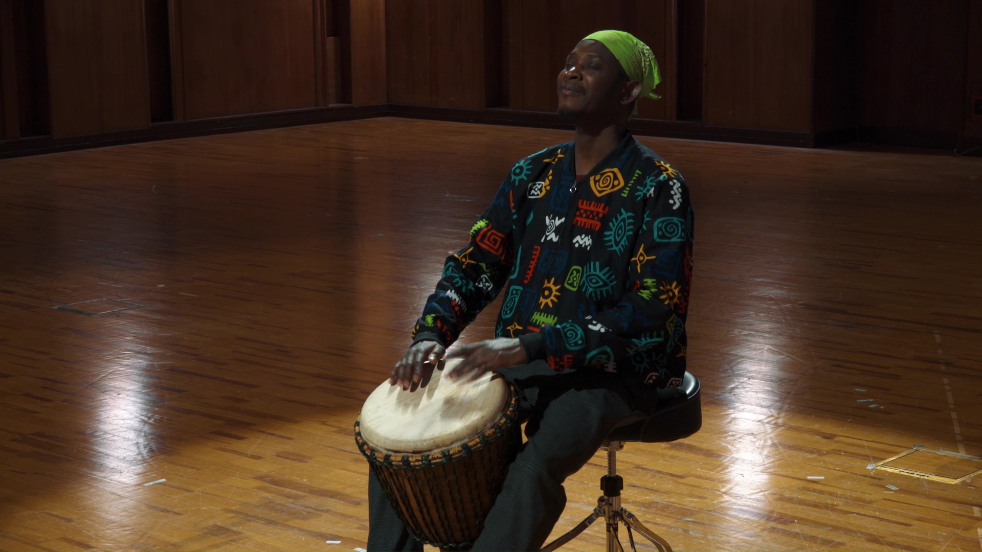 Man playing a djembe drum, wearing a colorful, culturally patterned jacket and lime green bandana, sitting on a stool on a polished wooden floor.