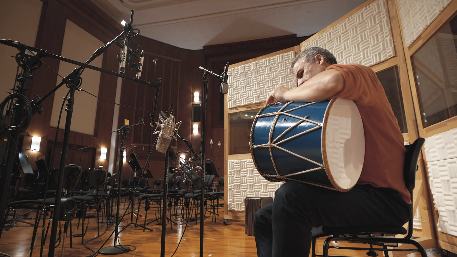 A musician playing a blue drum in a recording studio, surrounded by microphones and empty chairs set up for an orchestra.