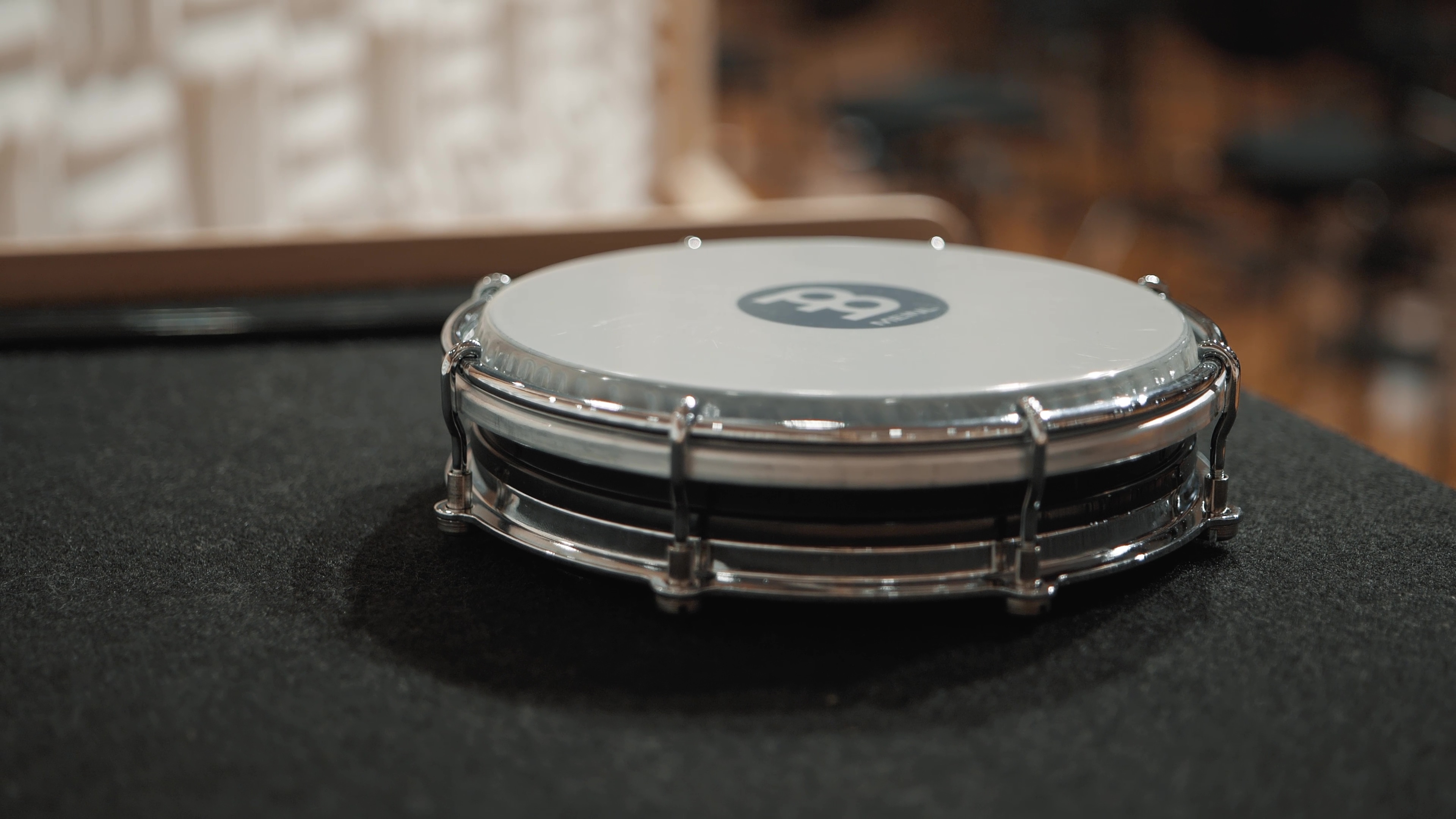 Top-down view of a Meinl snare drum with a white head, chrome hardware, and black drum shell, sitting on a dark grey textured surface in a music studio