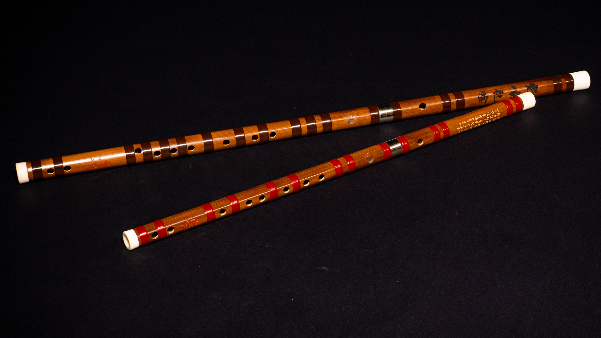 Two Chinese bamboo flutes, a darker wooden Dizi with brown bands and a lighter bamboo Xiao with red bands, resting diagonally on a black surface.