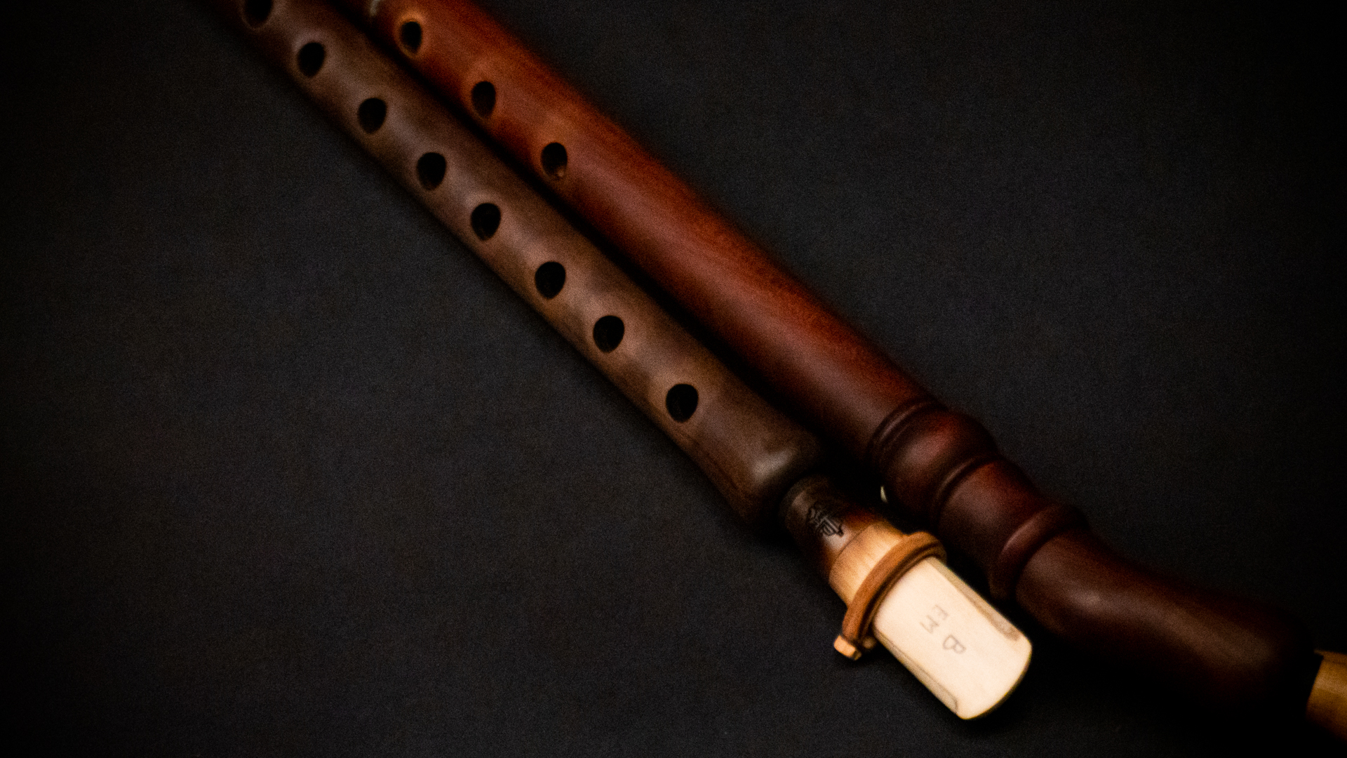 Two wooden wind instruments, possibly flutes or clarinets, with finger holes and a visible reed, resting on a dark surface.