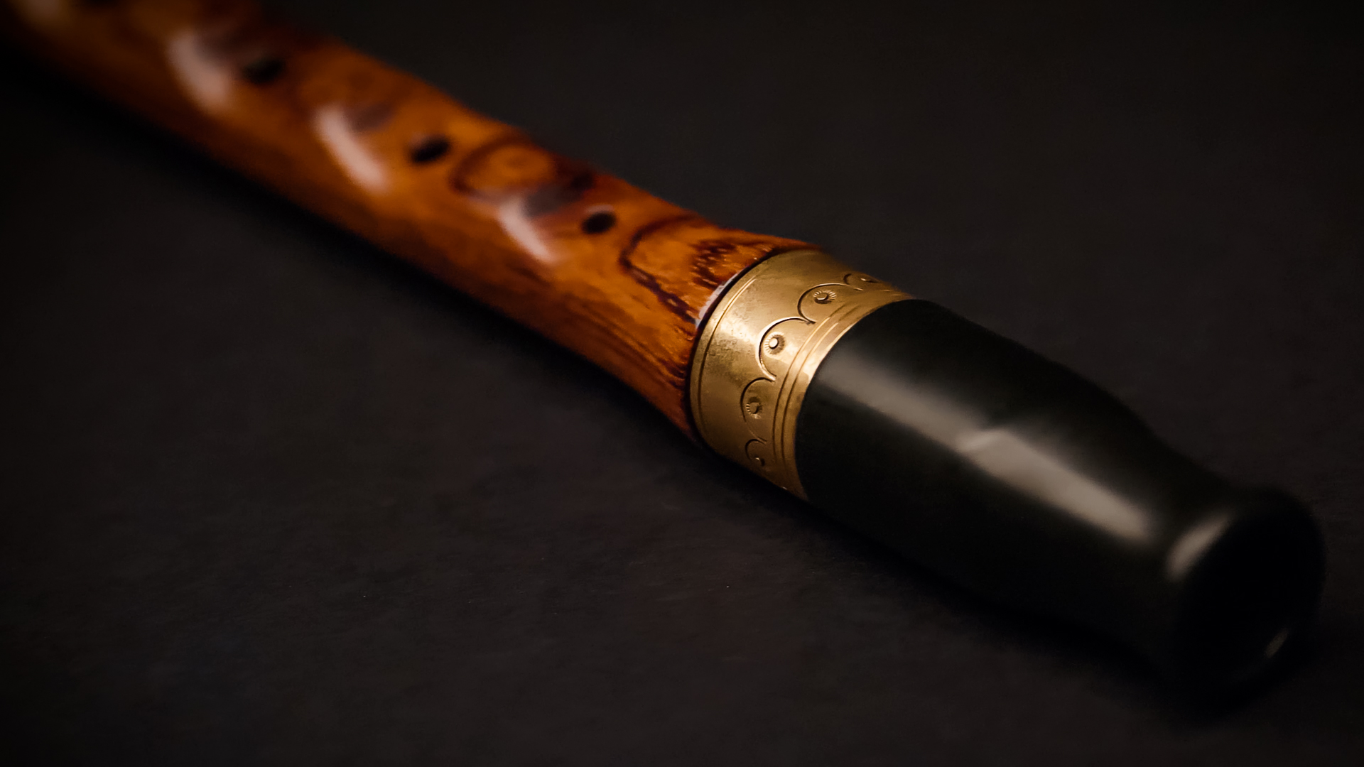 Close-up of a handcrafted wooden flute with a black mouthpiece and an ornate brass band, resting on a dark background.