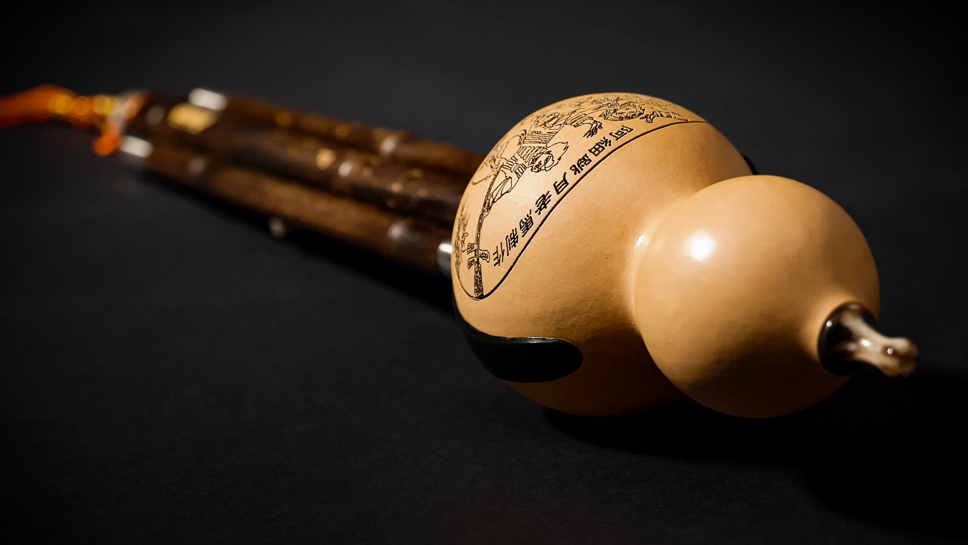 Close-up of a cucurbit flute or hulusi, a Chinese wind instrument with a light brown gourd resonator featuring etched traditional Asian artwork and dark wooden pipes extending into the background on a black surface.