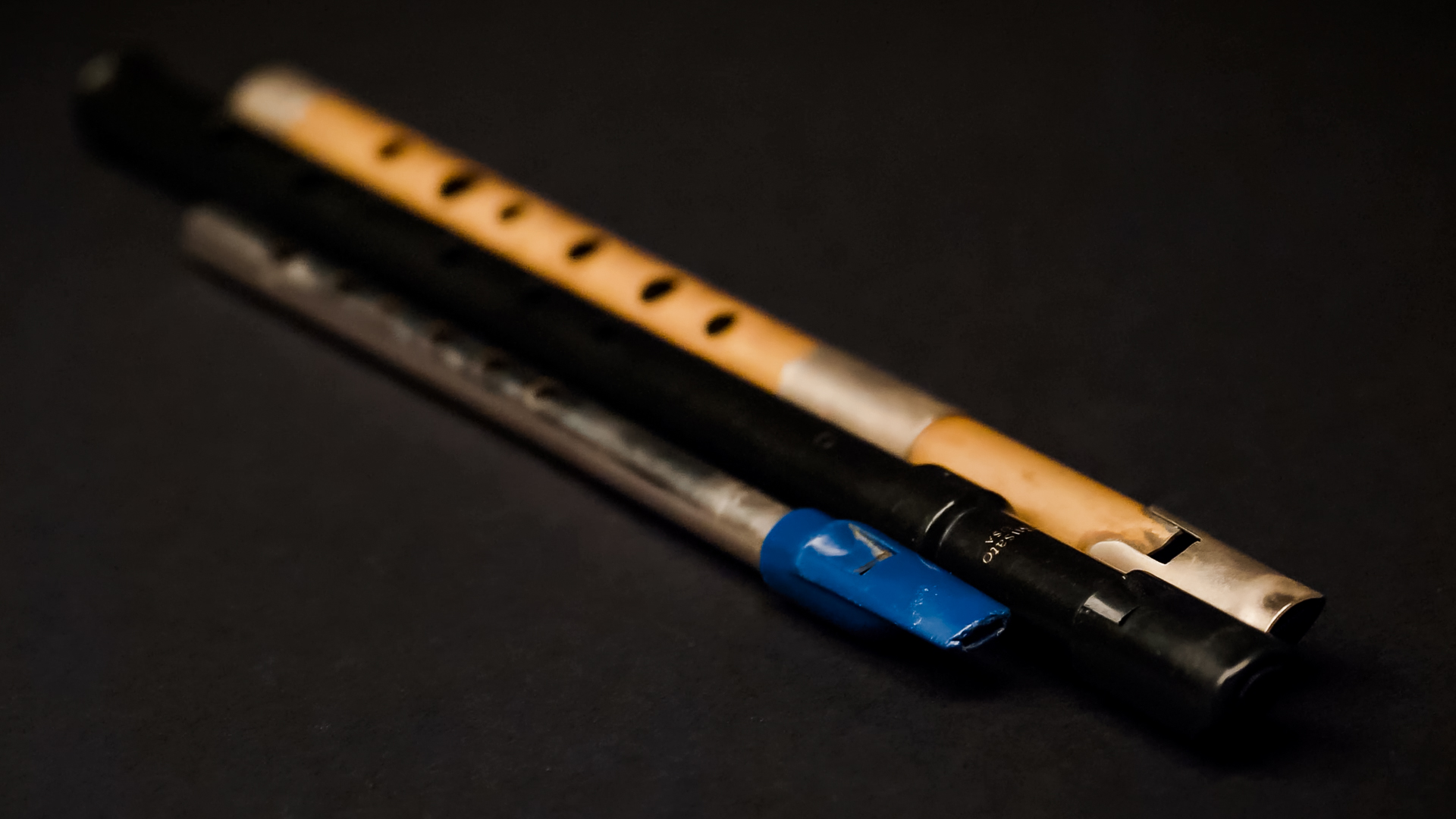 Three different woodwind instruments are lying diagonally on a dark surface. The top instrument is a light brown wooden flute with black finger holes and silver accents. Below it is a black flute, also with silver accents. The bottom instrument is a metallic silver flute with a bright blue mouthpiece.